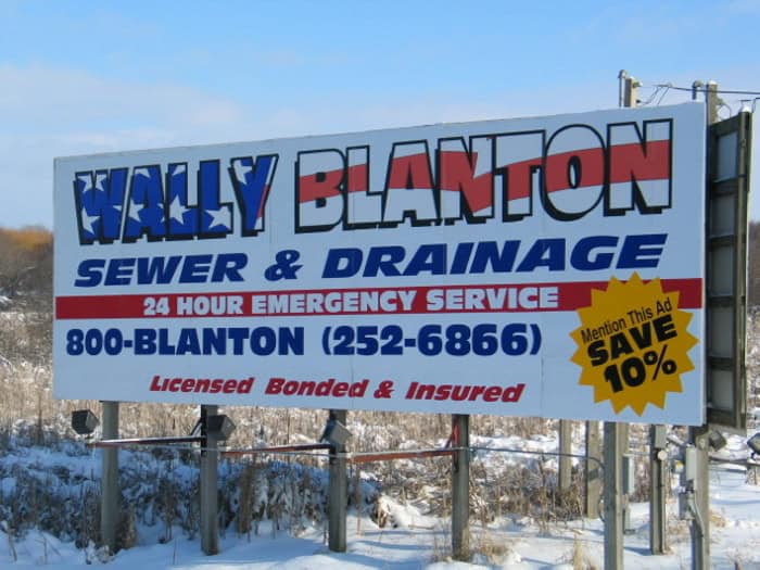 A roadside sign in snowy grass advertises "Wally Blanton Sewer & Drainage" with a 24-hour emergency service number and a 10% discount for mentioning the ad. Licensed, bonded, insured—and now offering vehicle wraps Spring Grove for your business.