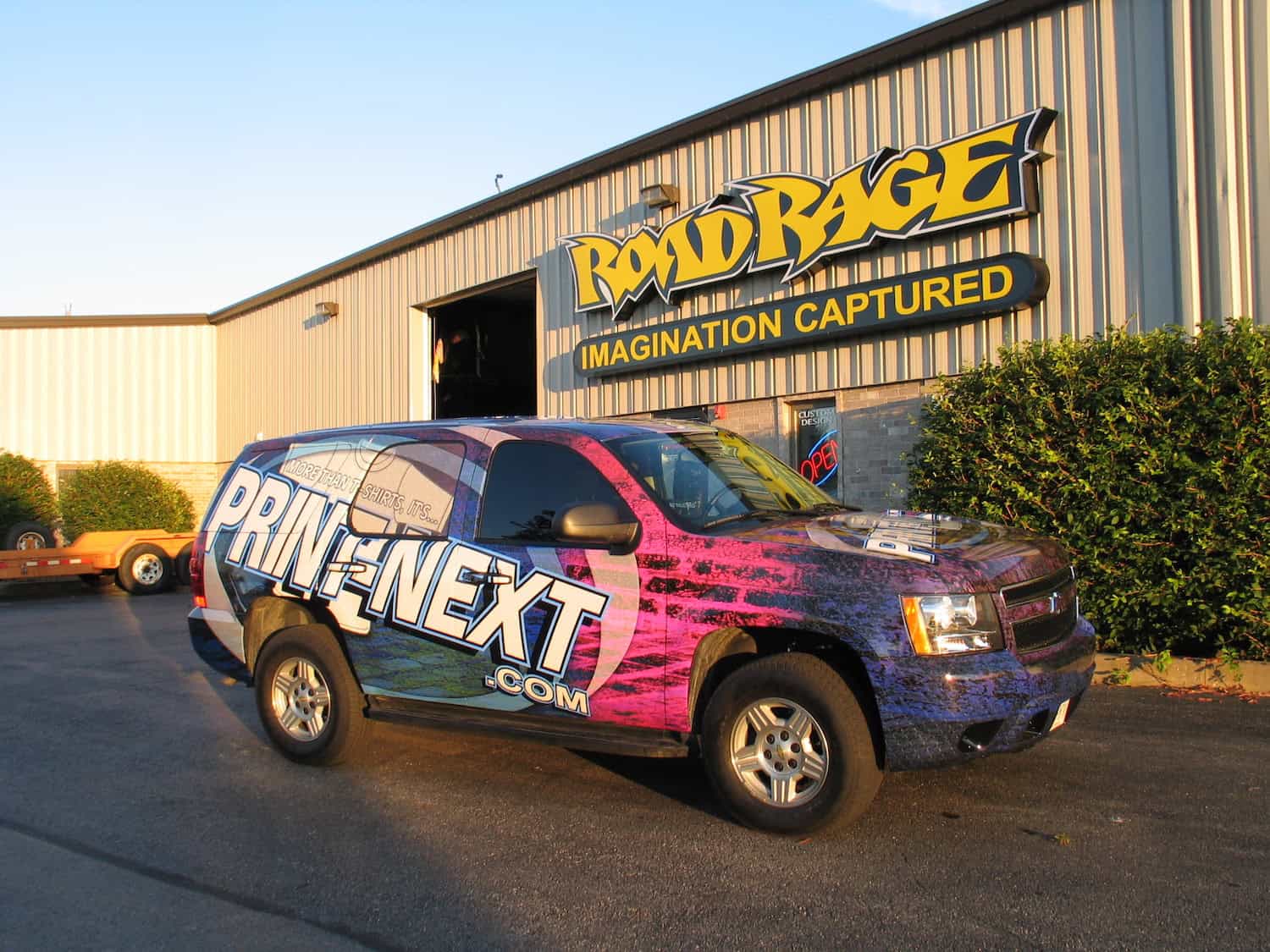 A large SUV with a colorful "PRINT-NEXT.com" wrap—showcasing premium vehicle wraps Spring Grove, IL—is parked in front of a building with a "ROAD RAGE IMAGINATION CAPTURED" sign above the entrance on a sunny day.