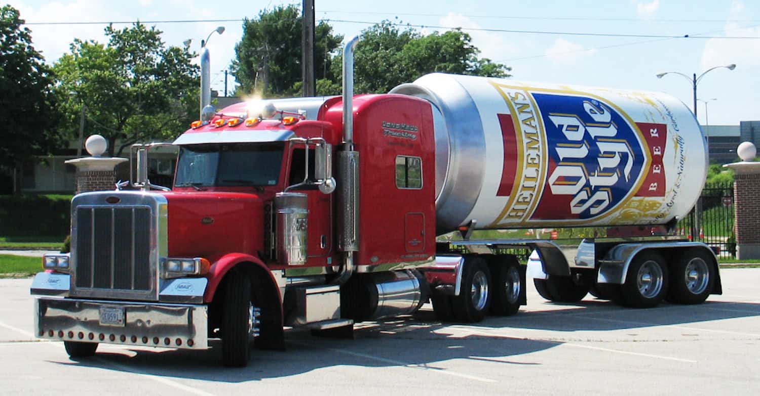 A red semi-truck parked in a lot in IL, hauling a trailer with a vehicle wrap designed to look like a giant can of Heileman's Old Style beer. Trees and a fence are visible in the background on a sunny day.
