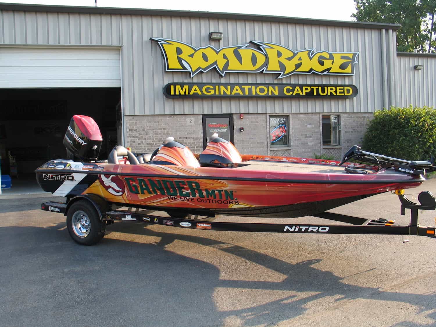 A red Nitro fishing boat with "Gander Mtn We Live Outdoors" graphics and custom vehicle wraps Spring Grove is parked on a trailer in front of a building with a "Road Rage Imagination Captured" sign in IL.