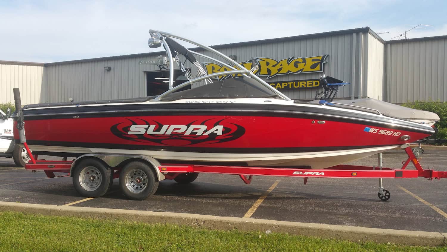 A red Supra speedboat with bold vehicle wraps Spring Grove graphics sits on a matching trailer in a parking lot, parked in front of a building with a "Rage Powersports" sign in IL.